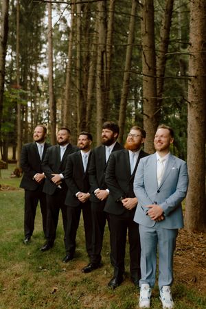 Groom in light blue suit stands with five groomsmen in black tuxedos among tall pine trees for wedding portrait.