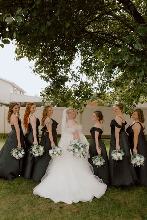 Bride in white gown poses with six bridesmaids in black dresses holding white and blue bouquets under large tree.