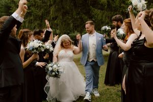 Bride and groom walk through cheering wedding party in black attire raising bouquets in outdoor celebration.