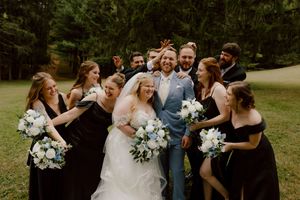 Joyful wedding party in black and light blue surrounds laughing bride and groom in lush green outdoor setting.