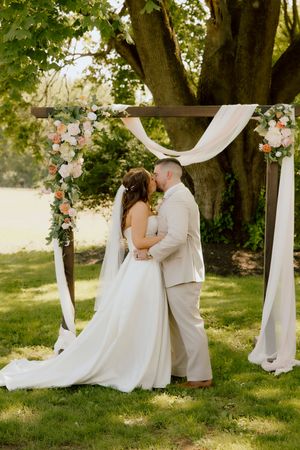Bride and groom share first kiss under floral wooden arch with white draping outdoors.