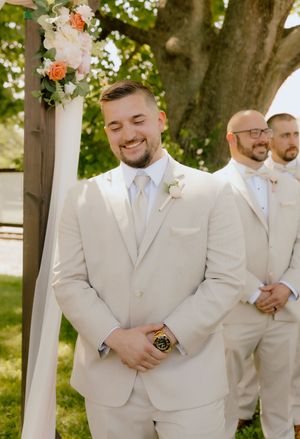 Handsome groom in cream suit with boutonniere smiles at altar during outdoor wedding ceremony.