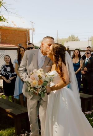Bride in white gown with peach bouquet shares first kiss with groom in grey suit at ceremony.