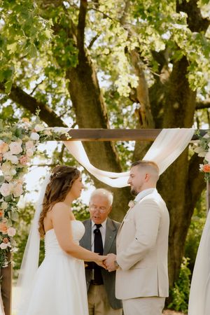Bride and groom hold hands at floral arch altar during outdoor wedding ceremony under large tree.