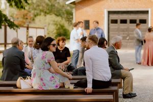 Wedding guests socializing on wooden benches at outdoor venue, woman in floral dress conversing with group.