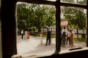 Wedding guests mingling in courtyard viewed through rustic window frame, surrounded by lush green trees.