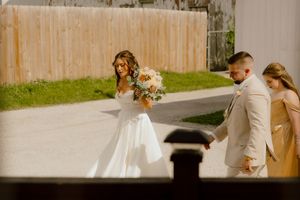 Bride in elegant white gown holding bouquet walks alongside groom in beige suit and bridesmaid in gold dress.