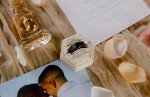 Wedding ring in hexagonal velvet box displayed with rose petals, perfume bottle, and couple photograph.