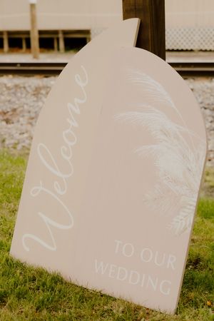 Elegant arched wedding welcome sign with white script lettering and floral design on blush background outdoors.