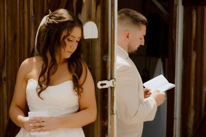 Bride in strapless white gown and groom in tan suit reading heartfelt vow letters beside a rustic wooden barn door.