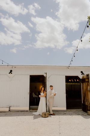 Bride and groom standing apart reading vows outside a white barn venue with string lights overhead under a blue sky.