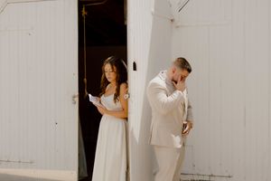 Emotional groom wiping tears while bride reads private vows on opposite sides of a white barn wall.