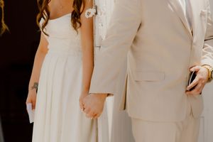 Close-up of bride and groom holding hands tenderly, bride in white gown with a floral tattoo on her wrist.