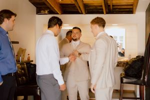 Groomsmen in cream and grey suits help each other with cufflinks while getting ready for wedding.