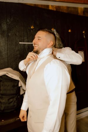 Smiling groom in cream vest and white shirt gets dressed with assistance before wedding ceremony.