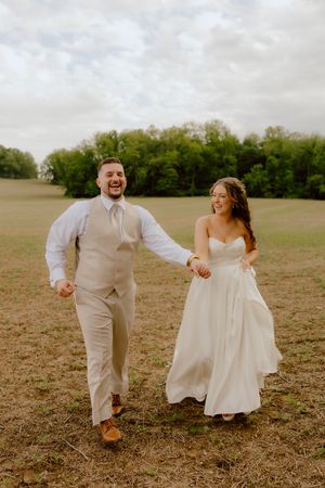 Laughing bride and groom run hand-in-hand through open field on wedding day.