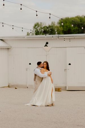 Groom in tan suit embracing and kissing bride in flowing white gown outdoors near a white barn with string lights.