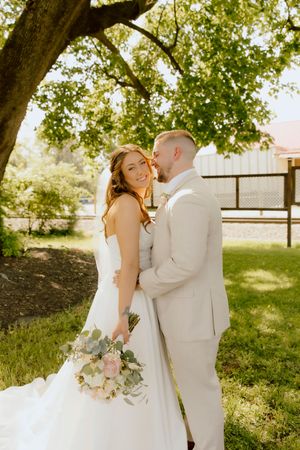Bride holding blush bouquet smiles as groom embraces her under large tree.