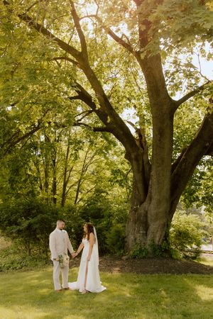 Bride and groom hold hands under a large sunlit tree in lush garden.