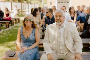 Couple in formal attire seated at outdoor wedding ceremony, woman in blue lace dress with floral sunglasses, man in beige suit.