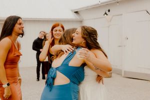 Bride hugs a guest in a blue dress while smiling women gather at an outdoor wedding celebration.