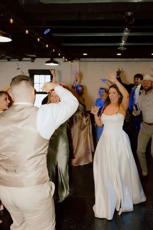 Bride in white gown dances energetically with guests celebrating on the dance floor at wedding reception.