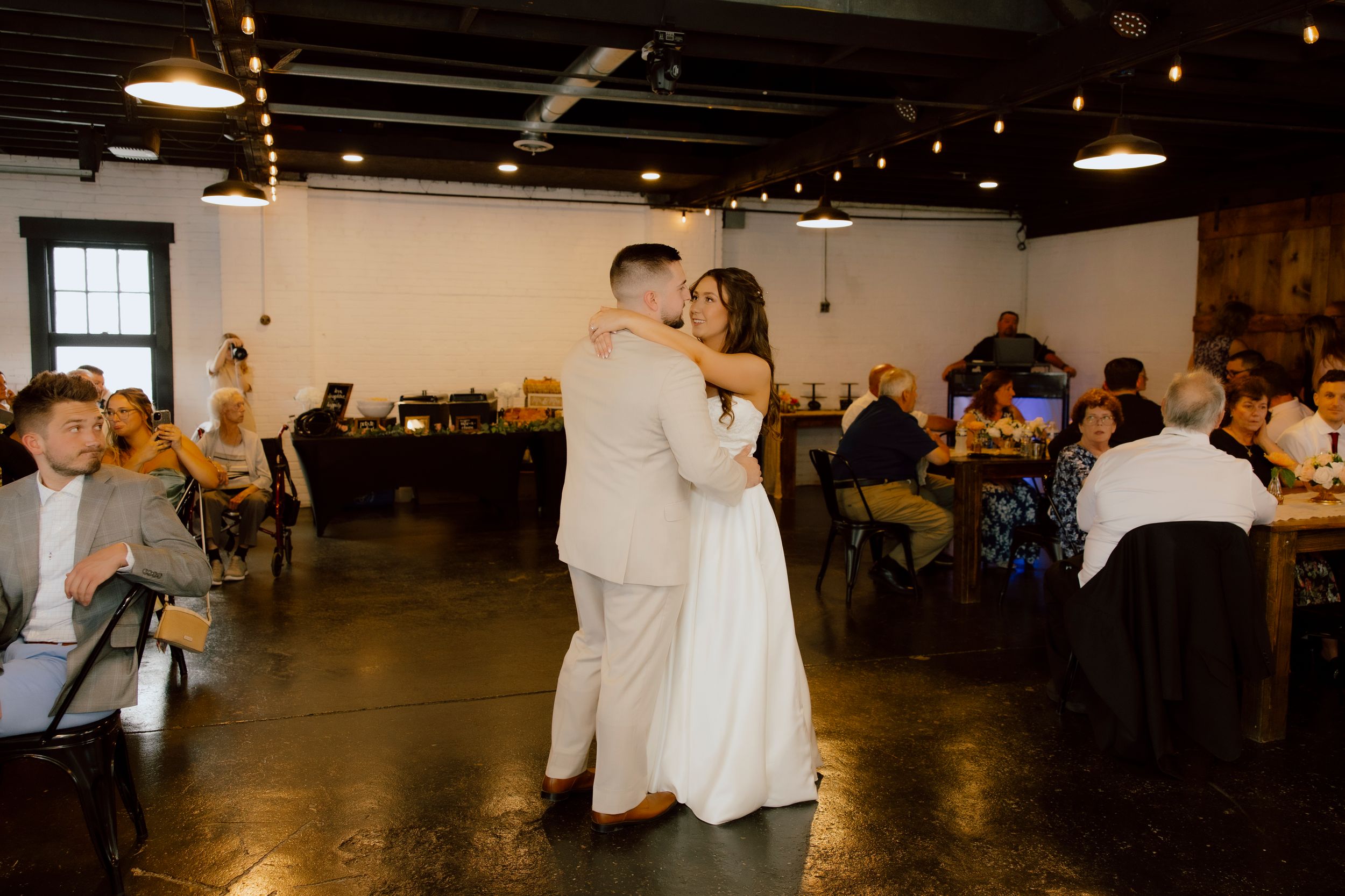 Bride and groom share their first dance in a warmly lit industrial wedding reception venue with guests seated.