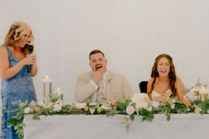Bride and groom laugh joyfully during a wedding speech at a floral-decorated sweetheart table.