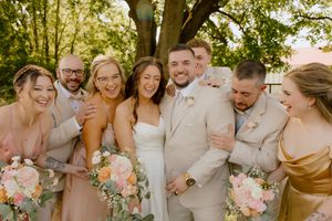 Joyful wedding party laughing together outdoors under a tree, bride in white gown surrounded by bridesmaids and groomsmen.