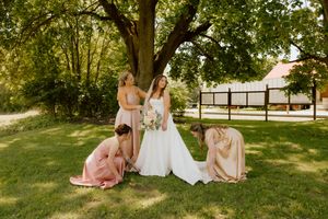 Bride holds bouquet while bridesmaids in pink satin dresses adjust her gown under a large tree outdoors.