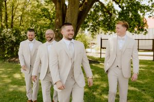 Groom and three groomsmen in matching beige suits walk confidently across a green lawn at a wedding.