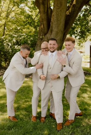 Groom laughs as three groomsmen in tan suits playfully pose together under a large tree outdoors.