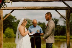 Bride and groom exchange vows under wooden arch officiated by minister near scenic pond.