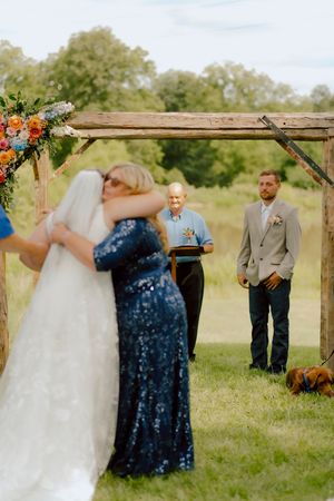 Mother embraces bride at altar while groom and officiant watch during outdoor wedding ceremony.