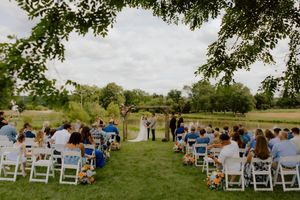 Outdoor wedding ceremony by a pond with guests seated in white chairs on green lawn.