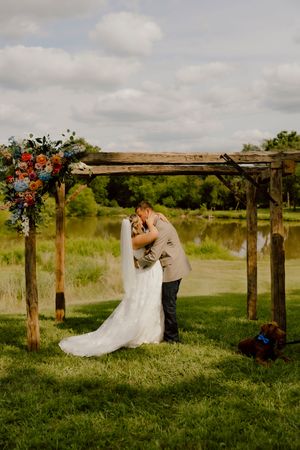 Bride and groom kiss under rustic wooden arch with colorful floral arrangements by a pond.