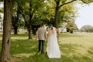 Bride and groom walk hand in hand through a tree-lined grassy field after wedding ceremony.