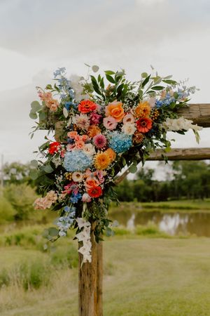 Colorful wedding arch adorned with orange, pink, blue, and white flowers with lush greenery near a pond.