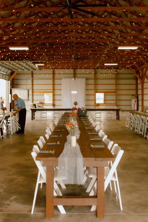 Elegant barn wedding reception with long wooden tables, white chairs, string lights, and floral centerpieces.