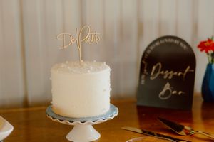 Simple white wedding cake with a gold DePatto cake topper displayed on a blue cake stand at a dessert bar.