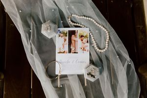 Wedding flat lay featuring a photo card, pearl necklace, rings, and a sheer veil on a dark surface.