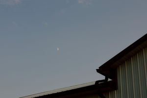 A crescent moon visible in the clear blue evening sky above the roofline of a metal barn building.