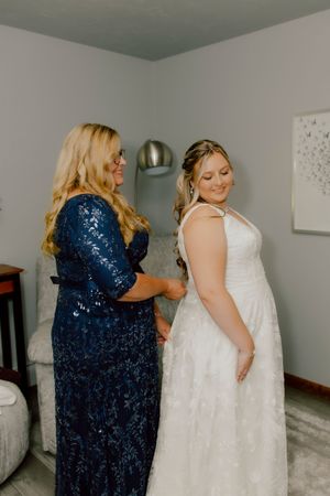 A bride in a white lace gown smiles as a woman in a navy sequin dress helps her prepare for her wedding day.