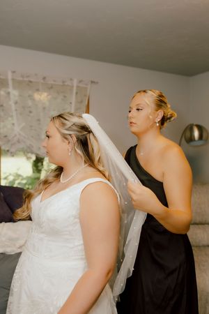 A bridesmaid in a black dress carefully adjusts the veil of a bride in a white lace wedding gown.