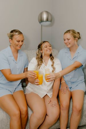Bride in white satin pajamas toasting orange juice with two bridesmaids in light blue matching sets while getting ready.