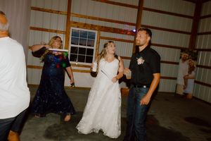 Bride in white gown dances joyfully with groom and guests under string lights at a barn wedding reception.