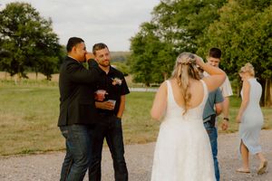 Wedding guests socializing outdoors on a gravel path surrounded by green fields and trees.