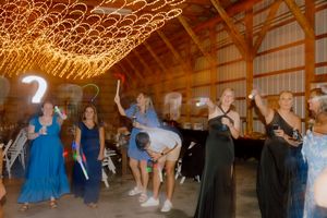 Wedding reception guests dance and wave light sticks under glowing string lights inside a rustic barn venue.