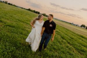 Bride and groom walk hand-in-hand through a lush green field at sunset during their wedding day.