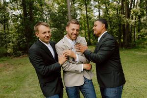 Groom laughs joyfully as two groomsmen playfully embrace him in a lush green outdoor setting.